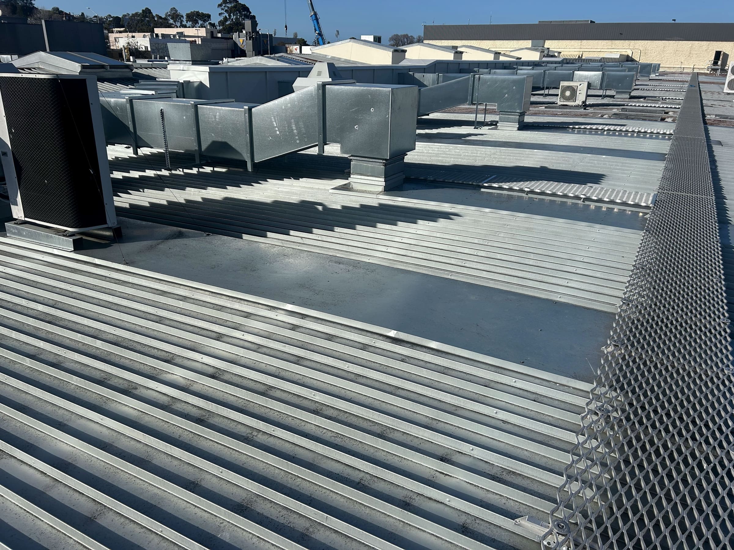 An existing commercial metal roof with large ducting runs, condenser units and walkway mesh.